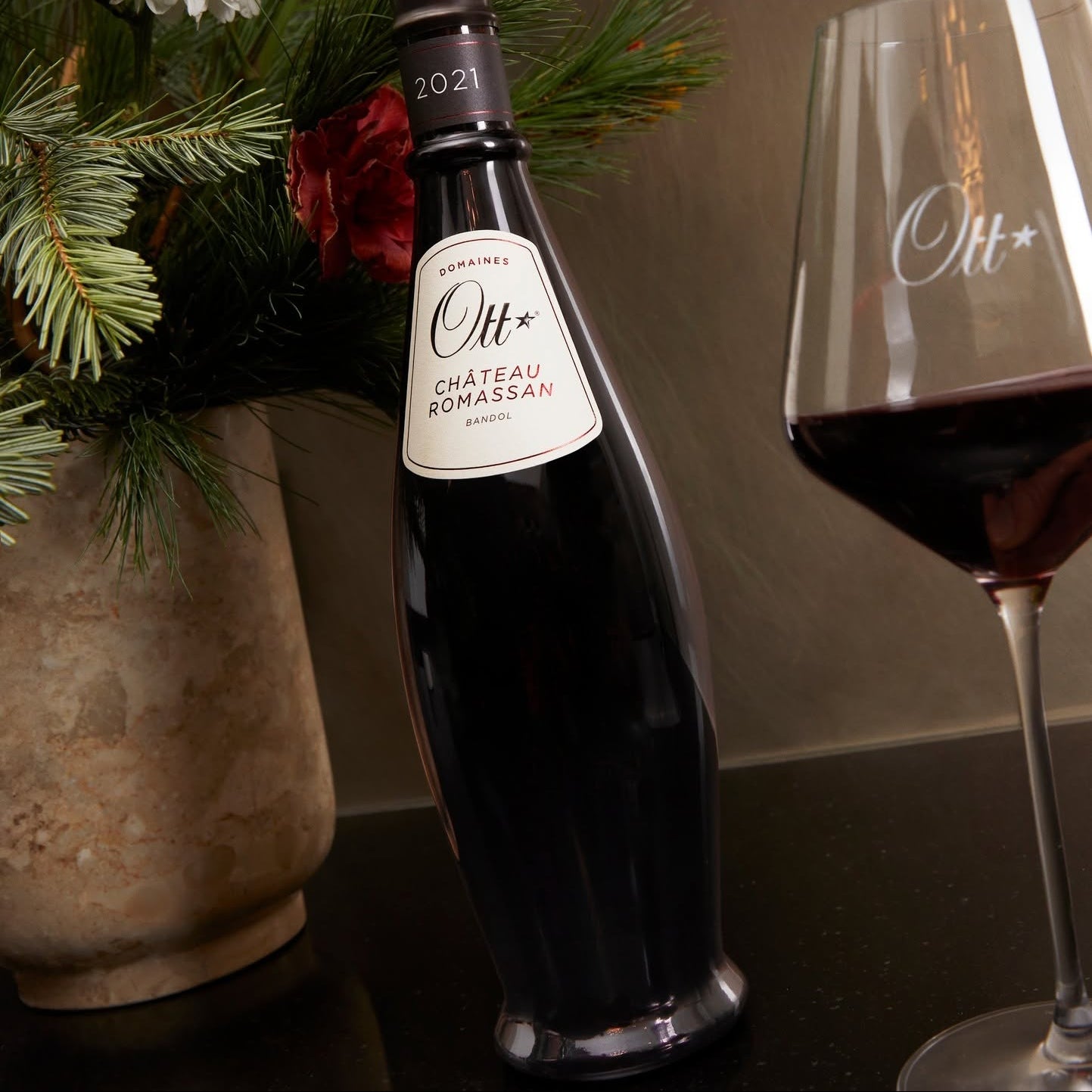Load image into Gallery viewer, A bottle of Chateau Romassan Bandol red wine stands on a dark tabletop beside a filled wine glass, with a floral arrangement in the background.