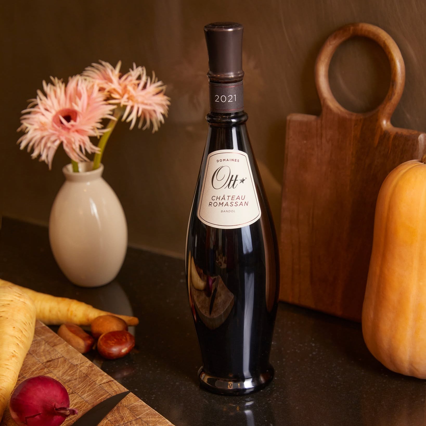 Load image into Gallery viewer, A bottle of Château Romassan Ott Bandol red wine stands on a dark countertop in a kitchen setting, surrounded by a small vase of pink flowers, a cutting board with a radish and chestnuts, and a yellow squash.