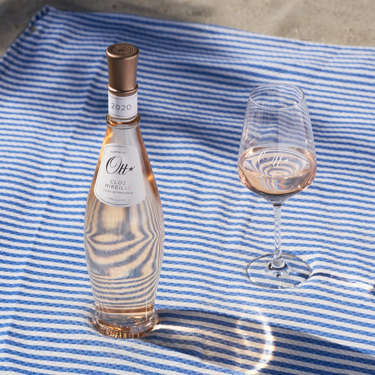 Load image into Gallery viewer, A bottle of Ott Mireille Rosé wine and a matching wine glass filled with rosé wine, placed on a blue and white striped blanket outdoors on sand with dappled sunlight.