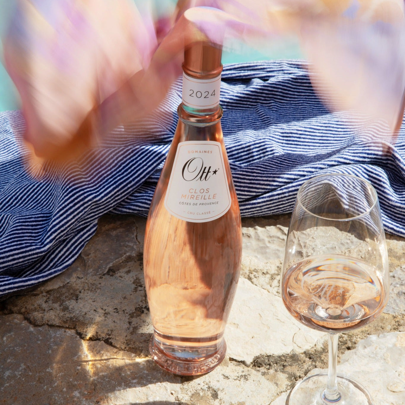 Load image into Gallery viewer, Ott Clos Mireille Cotes de Provence Rose wine bottle and glass on a stone surface with blurred colourful fabric in the background