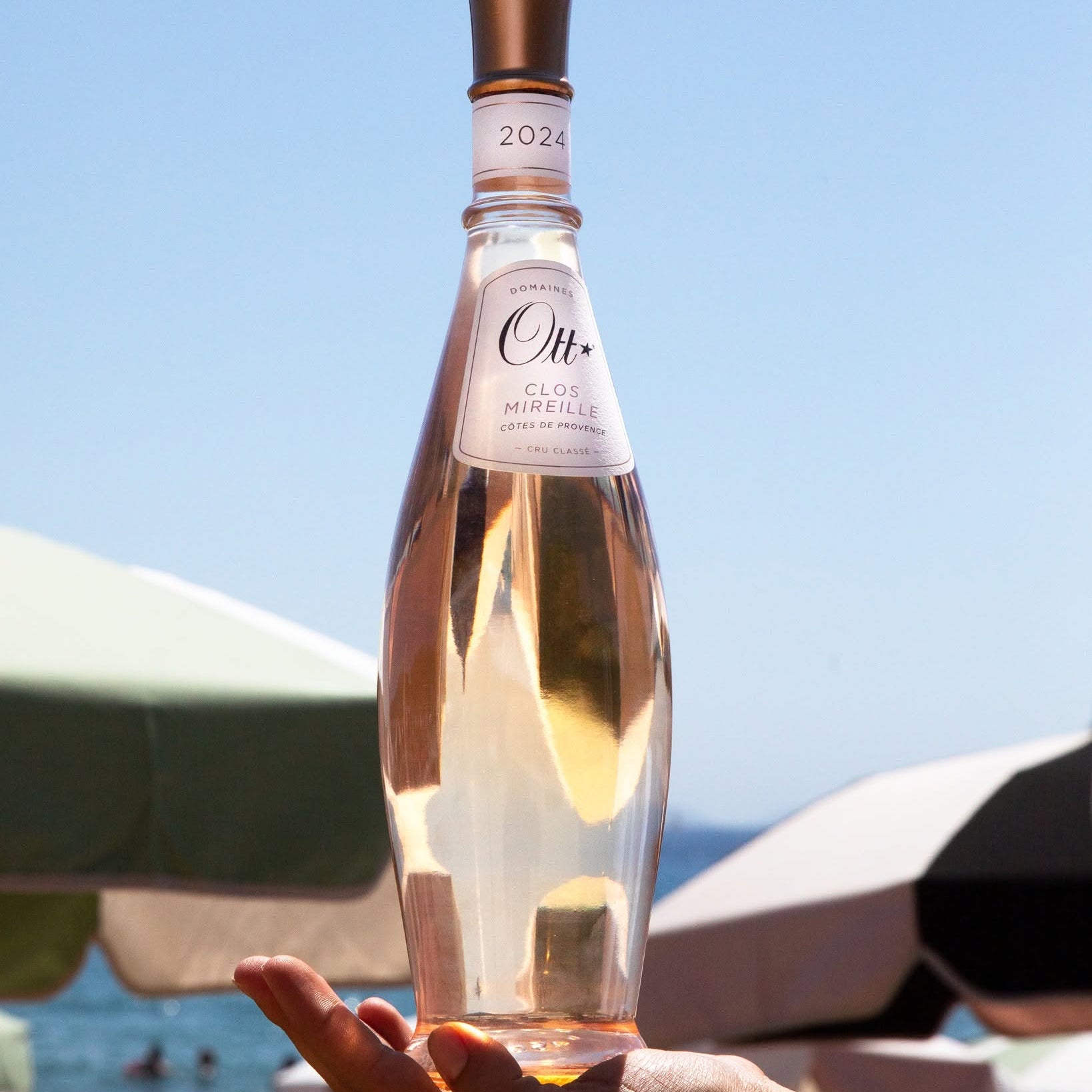 Load image into Gallery viewer, A hand holds a tall glass bottle of Ott Clos Mirielle rosé wine against a sunny beach background with umbrellas and the sea.