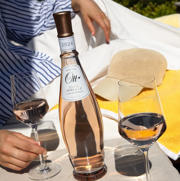 Load image into Gallery viewer, A bottle of Ott Mireille rosé wine with two filled wine glasses, set on a table outdoors with a person's hand and arm visible, along with a straw hat and white clothing in the background.