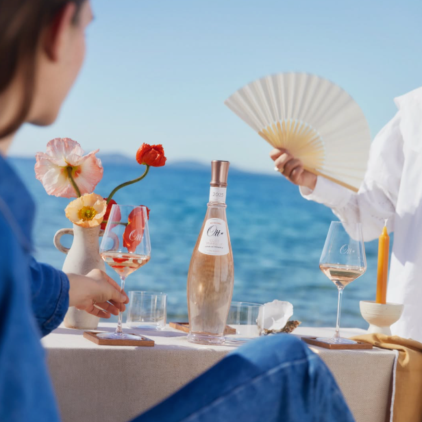 Load image into Gallery viewer, A lifestyle scene by the ocean showing a table set with a bottle of Ott Mirielle rosé wine, glasses, flowers, and a folding fan, with two people partially visible in a bright outdoor setting.