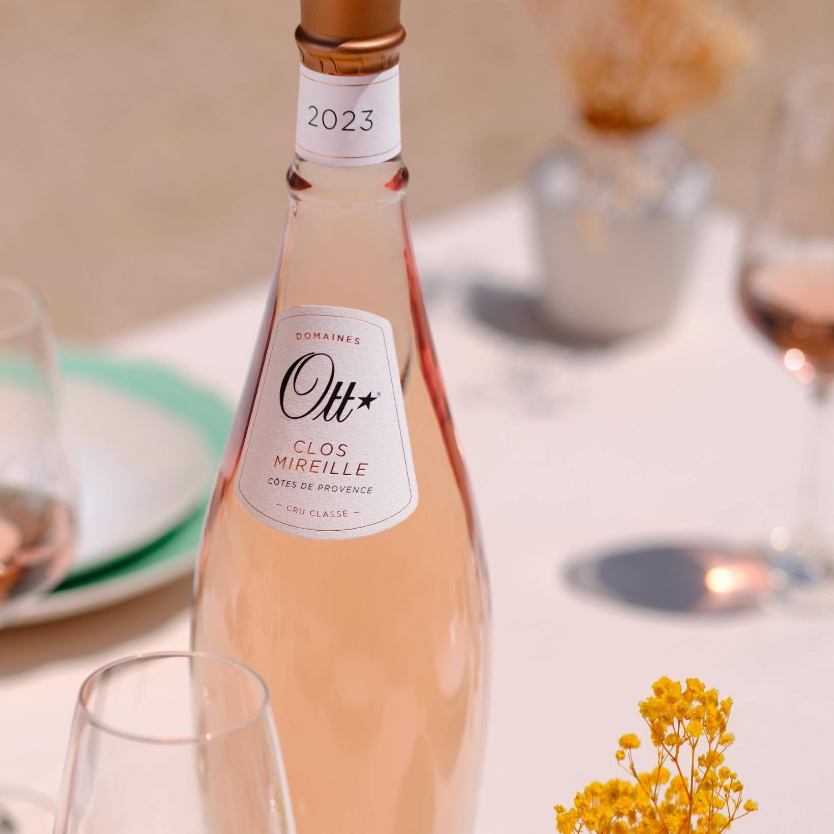 Load image into Gallery viewer, A bottle of Clos Mirelle Rosé rosé wine with a gold foil top and white label, set on a white table with wine glasses and dried flowers in a softly blurred outdoor setting.
