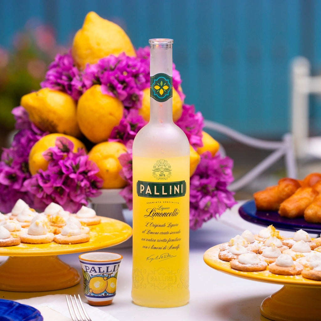Pallini limoncello bottle on a table with lemons, flowers, and pastries.