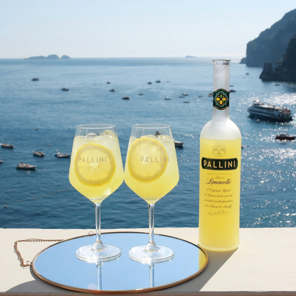 Two glasses of limoncello with a bottle on a reflective surface with a scenic ocean view.
