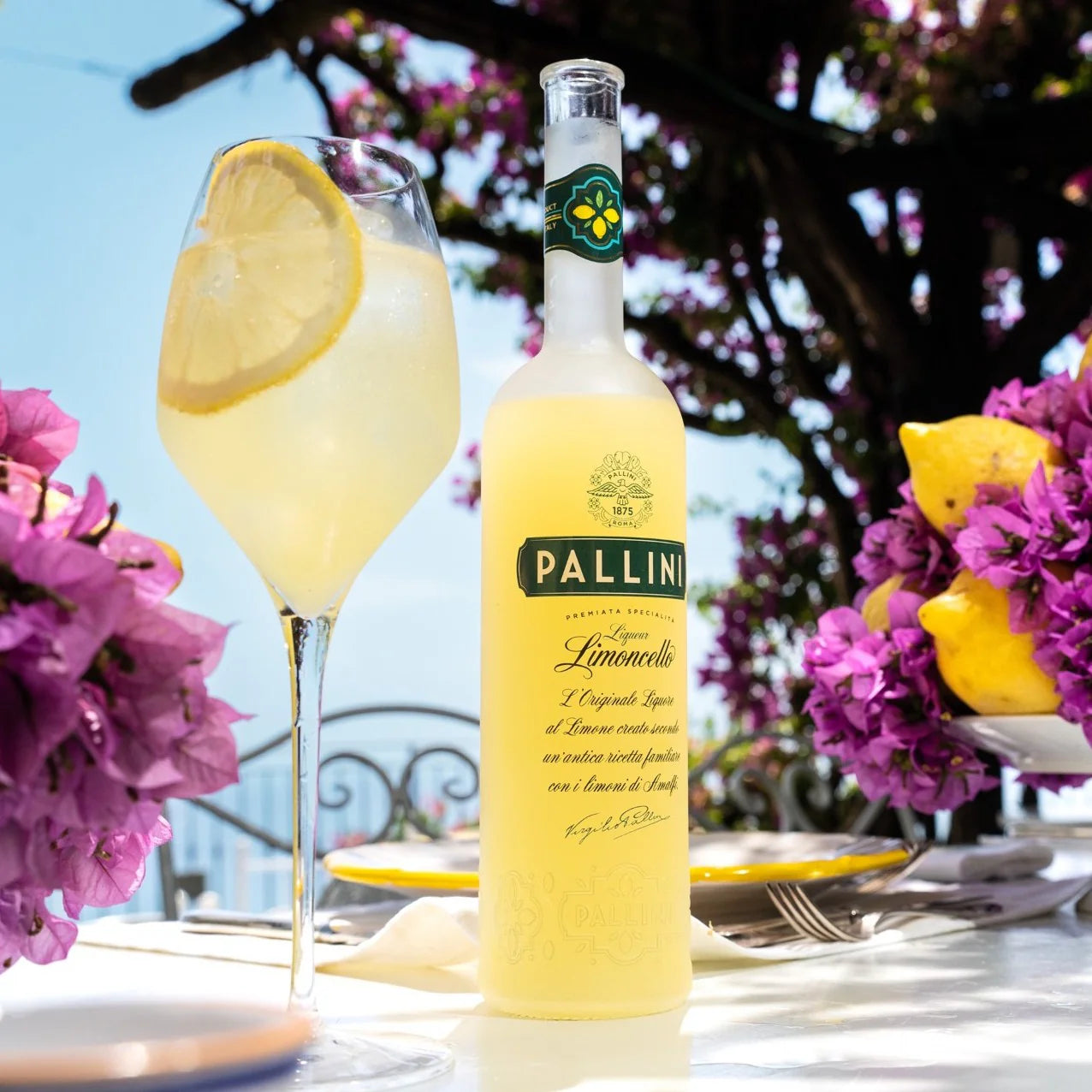 Load image into Gallery viewer, Pallini Limoncello bottle with a glass of limoncello on a table with flowers and lemon slices.