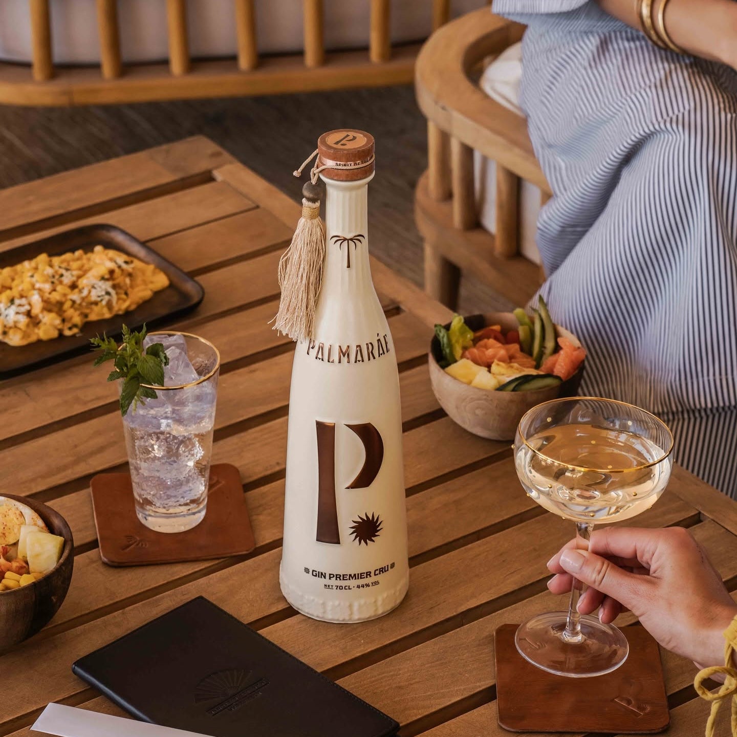 Load image into Gallery viewer, A bottle of Palmaráe Gin Premier Cru is displayed on a wooden table in a restaurant setting, surrounded by a glass of water, a cocktail glass with a drink, and a hand holding a cocktail glass.
