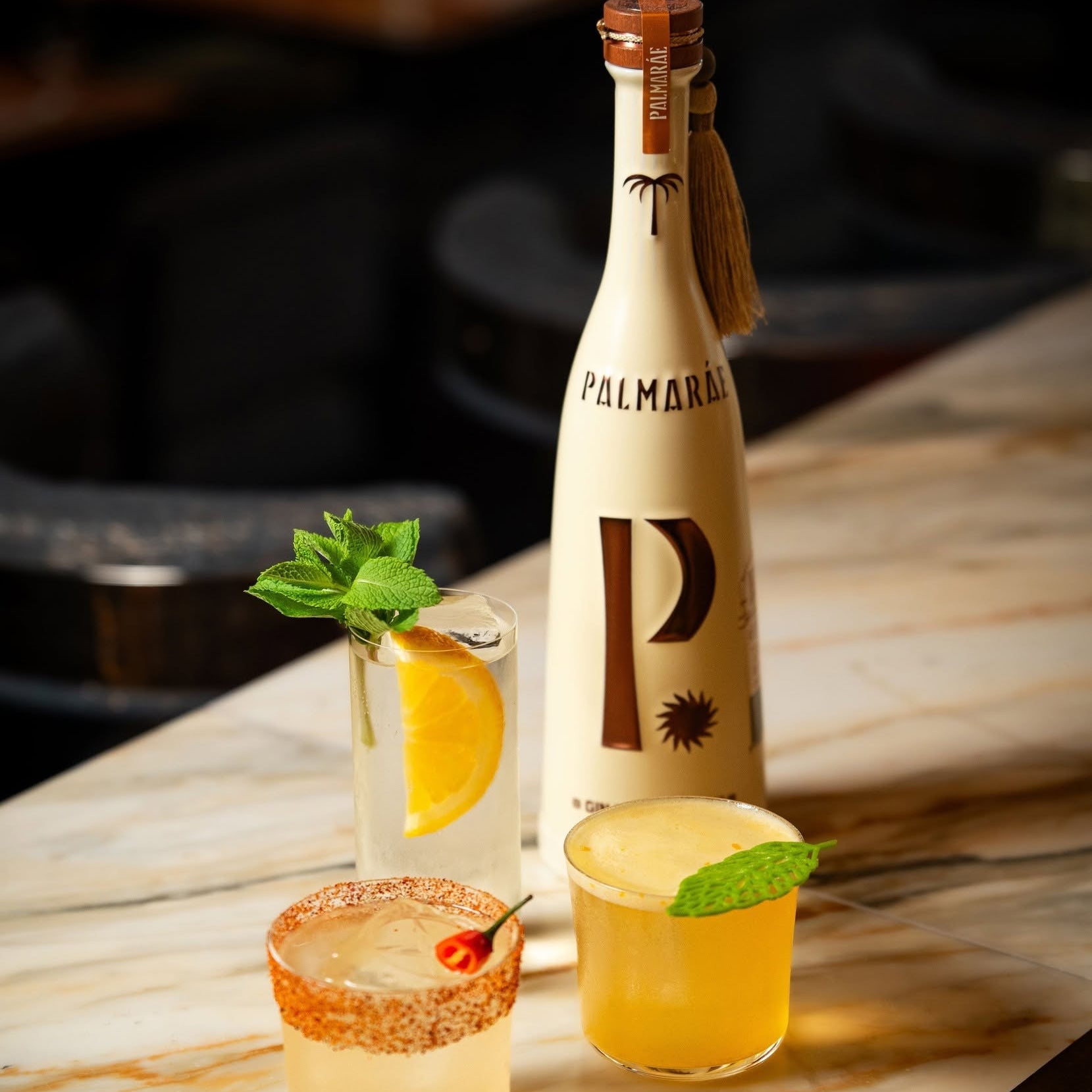 Load image into Gallery viewer, A bottle of Palmaráe Gin Premier Cru stands on a light marble bar surface beside three prepared cocktails: a margarita with a salted rim, a yellow cocktail with a lime wedge, and a pale yellow cocktail with a mint garnish.