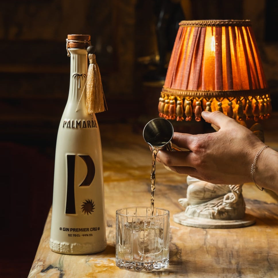 Load image into Gallery viewer, A bottle of Palmaráe Gin Premier Cru is shown on a marble table beside a lit decorative lamp, with a hand pouring liquid from a small container into a glass.