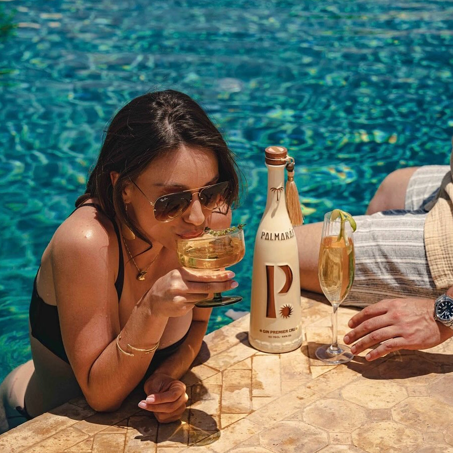 Load image into Gallery viewer, A woman in a black swimsuit and sunglasses leans over a swimming pool holding a cocktail, next to a bottle of Palmaráe Gin Premier Cru and a man's hand at the poolside.