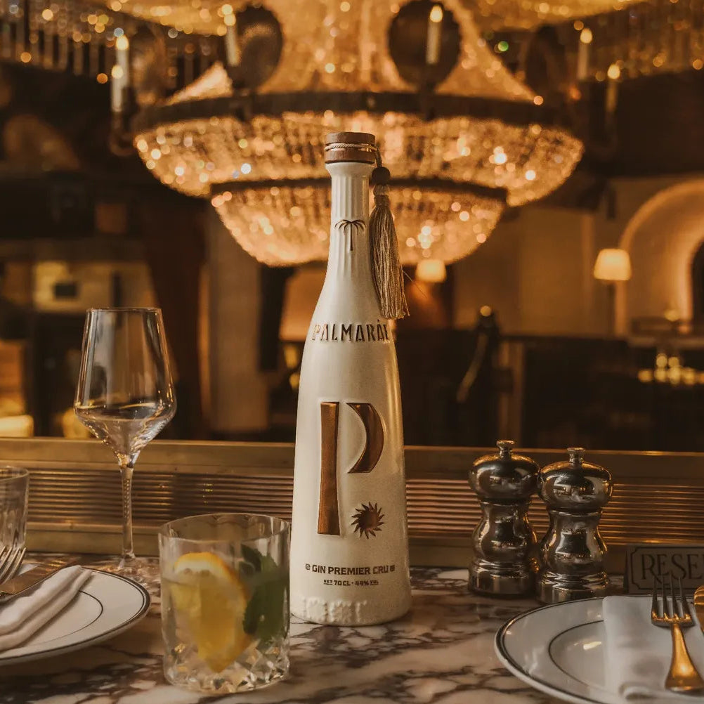 Load image into Gallery viewer, A bottle of Palmaráe Gin Premier Cru sits on a marble table set with plates, a wine glass, a gin and tonic drink garnished with lemon and lime slices, under a warm, glowing chandelier.