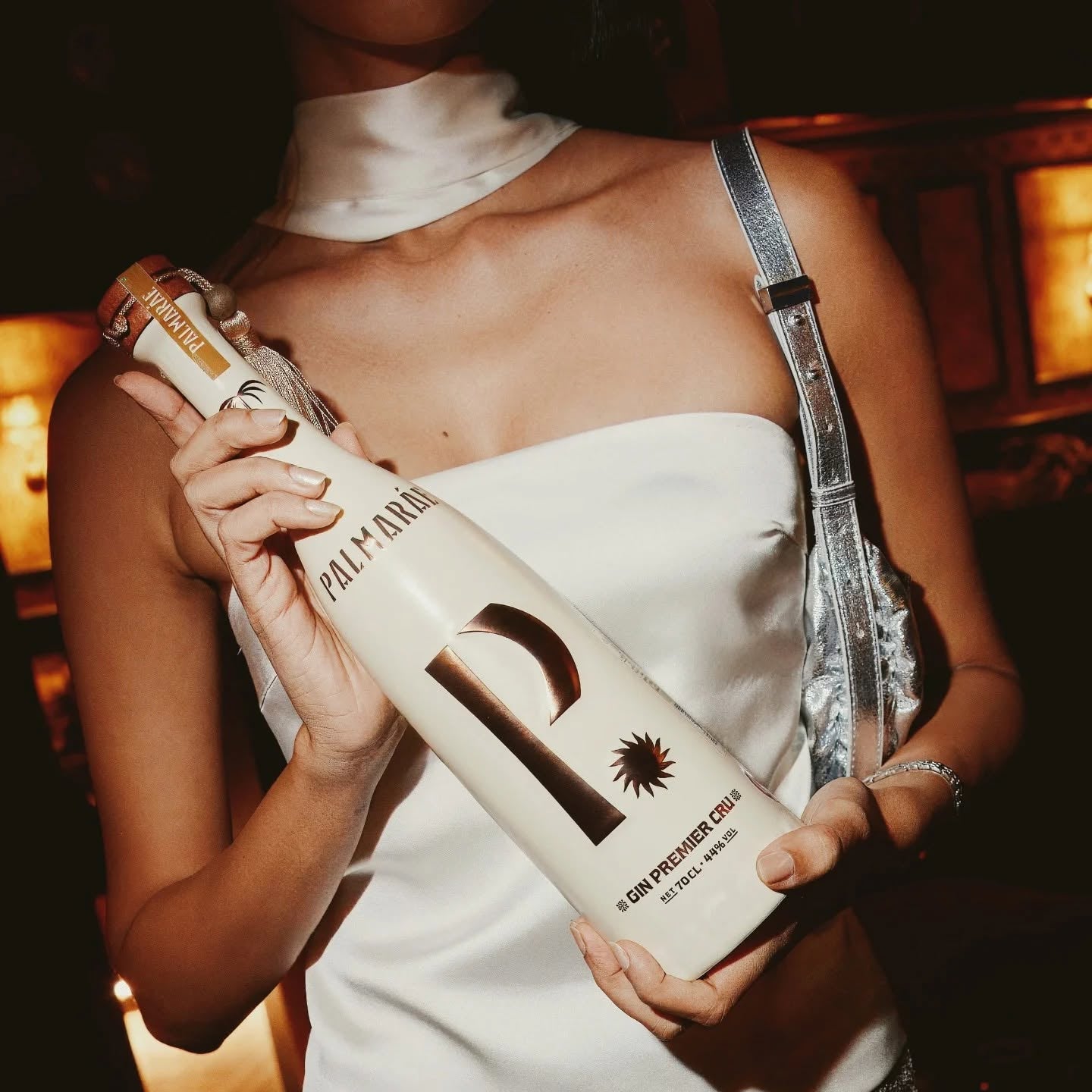 Load image into Gallery viewer, A stylish woman dressed in a white strapless top and silver sequinned skirt holds a bottle of Palmaráe Gin Premier Cru in a chic restaurant or bar setting.