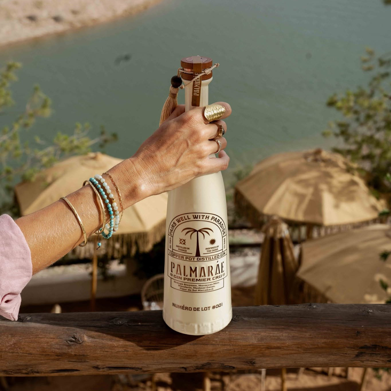 Load image into Gallery viewer, A woman holds a bottle of Palmaráe Gin Premier Cru against an outdoor patio backdrop with umbrellas and a lake in the background.