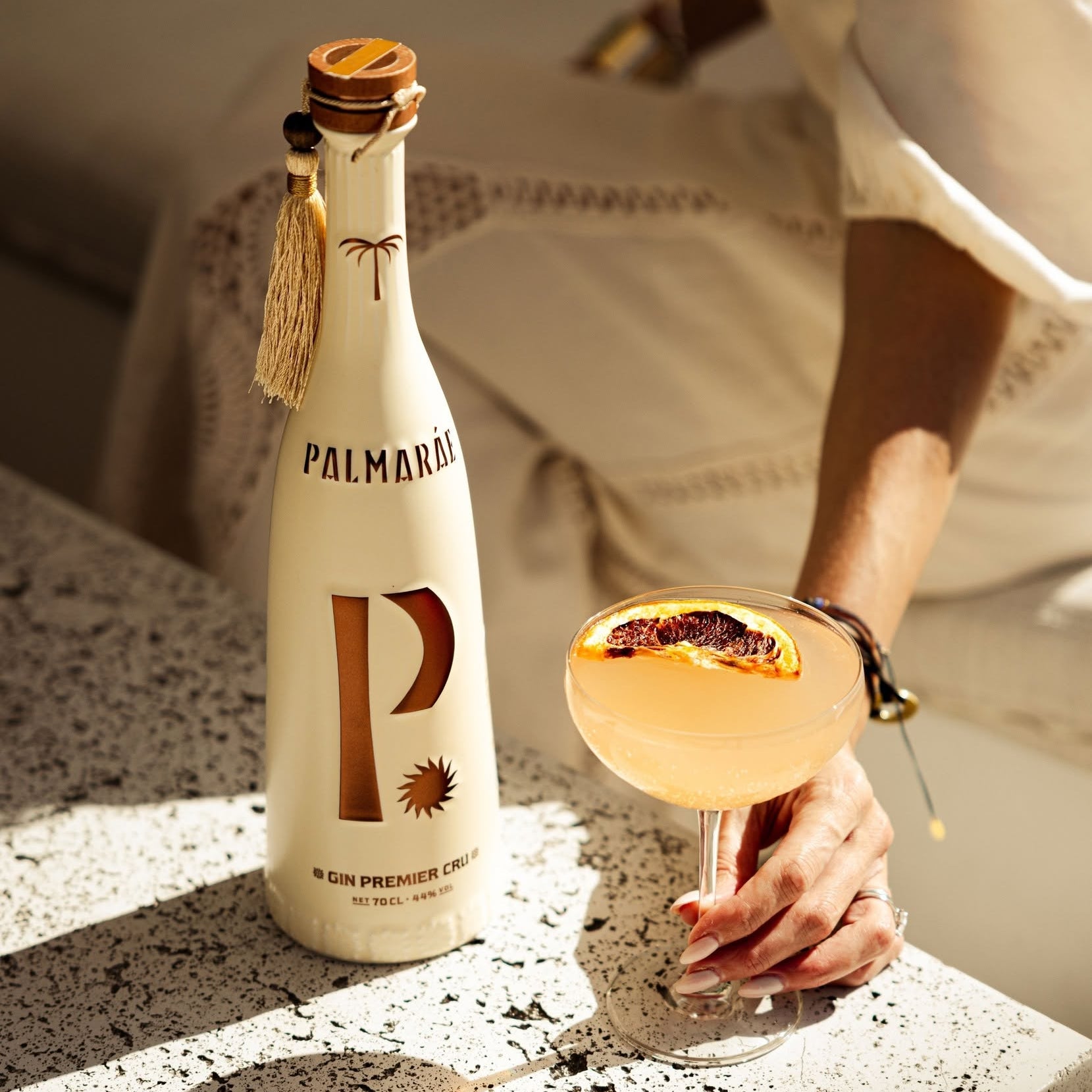 Load image into Gallery viewer, A bottle of Palmaráe Gin Premier Cru stands on a speckled stone surface beside a coupe glass filled with a pale orange cocktail garnished with citrus, with a woman's hand and torso visible in a warm indoor setting.