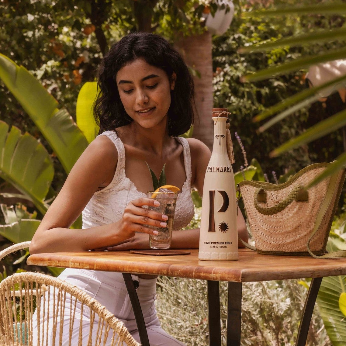 Load image into Gallery viewer, A woman sits at a small outdoor table surrounded by lush greenery, holding a cocktail. On the table is a bottle of Palmaráe Gin Premier Cru and a woven straw tote bag.