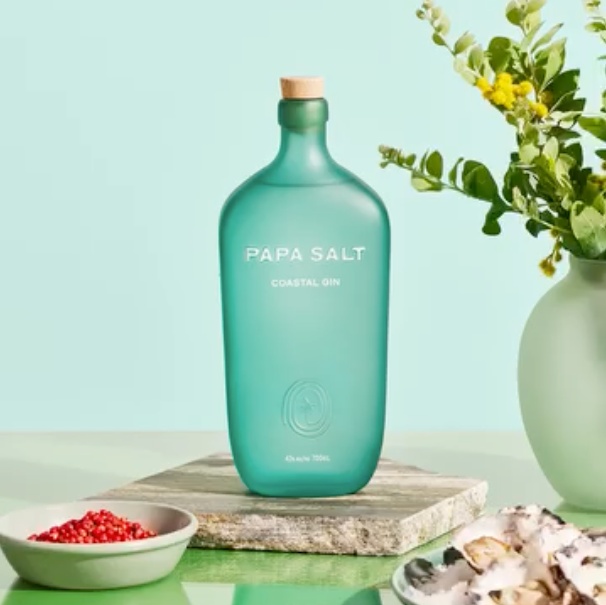 Load image into Gallery viewer, A bottle of Papa Salt Coastal Gin next to Pomegranate seeds and plant, with a teal background