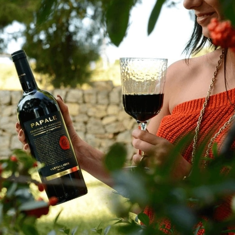 Load image into Gallery viewer, A woman holding a bottle of Papale Oro Varvaglione Primitivo di Manduria red wine in the garden with a glass in the other hand