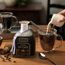 Load image into Gallery viewer, Person pouring milk into a glass of coffee with a bottle of Patron XO Cafe liqueur on a wooden table.
