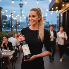 Load image into Gallery viewer, Woman holding a bottle of Pegasus Bay Sauvignon Blanc white wine in an outdoor evening setting with string lights and people in the background.