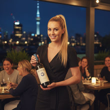 Load image into Gallery viewer, Woman holding a bottle of Pegasus Bay Sauvignon Semillon Bordeaux-style blend white wine with a city skyline in the background
