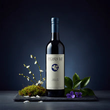 Load image into Gallery viewer, a bottle of red wine on a table with some flowers on a blue back ground.
