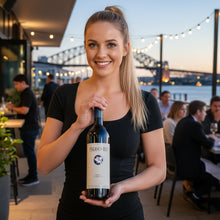 Load image into Gallery viewer, Waitress holding a bottle of Pegasus Bay merlot cabernet red blend wine outdoors with a bridge and people in the background