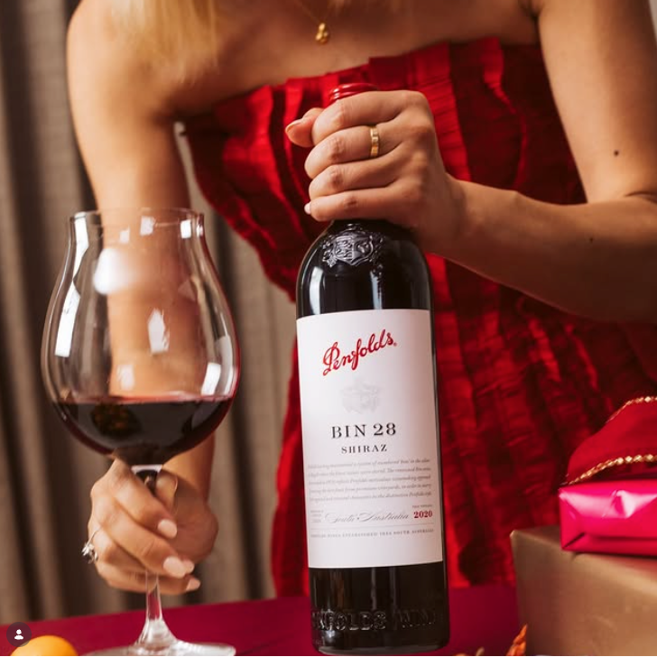 Load image into Gallery viewer, A woman in a red dress holds a bottle of Penfolds Bin 28 Shiraz red wine next to a filled wine glass, with wrapped gifts in the background.