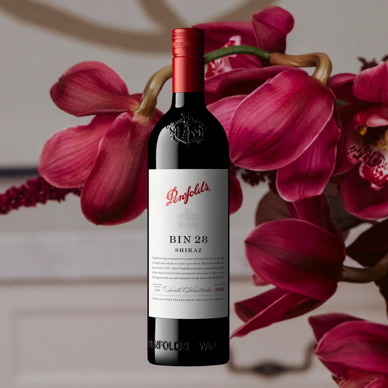 Load image into Gallery viewer, A bottle of Penfolds Bin 28 Shiraz red wine centred against a soft-focus background with large pink orchids. White text at the top reads 'BIN 28 SHIRAZ' and a descriptive text block appears along the bottom.