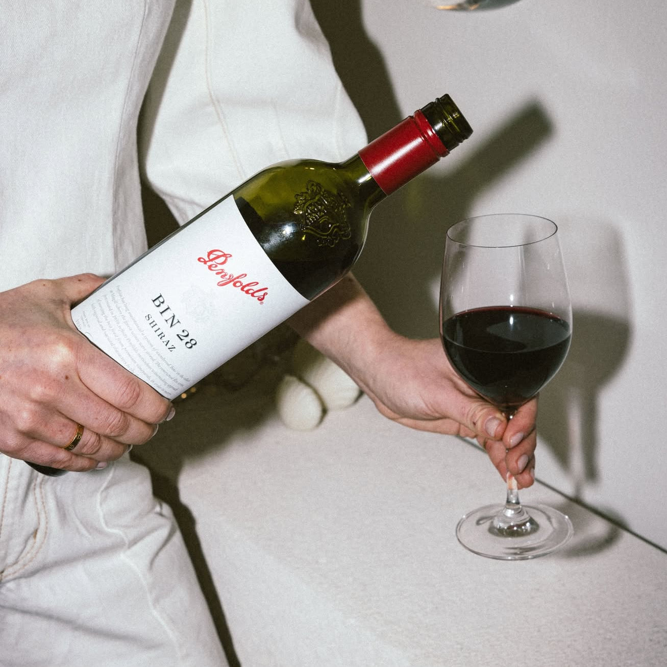 Load image into Gallery viewer, A person in a white outfit holds a bottle of Penfolds Bin 28 Shiraz red wine and a glass of red wine in a dimly lit interior setting.