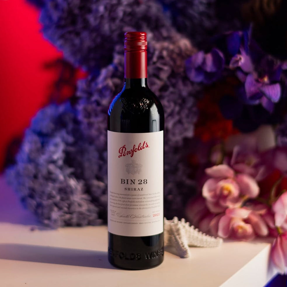Load image into Gallery viewer, A bottle of Penfolds Bin 28 Shiraz wine stands on a white ledge in front of a colourful floral arrangement with red, blue, and purple flowers under dramatic coloured lighting.