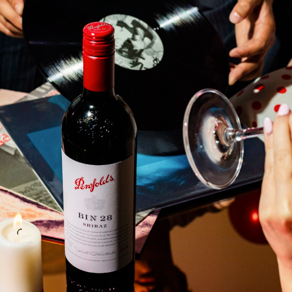 Load image into Gallery viewer, A bottle of Penfolds Bin 28 Shiraz red wine stands on a table in a dimly lit, cozy setting with a lit candle, a vinyl record, and a wine glass nearby.
