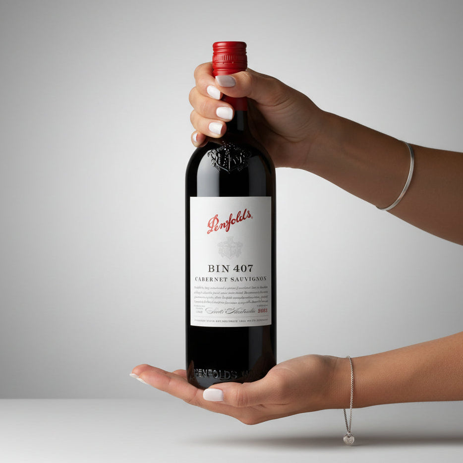 Load image into Gallery viewer, A hand holds a bottle of Penfolds Bin 407 Cabernet Sauvignon, with the other hand supporting the base, against a clean grey studio background.