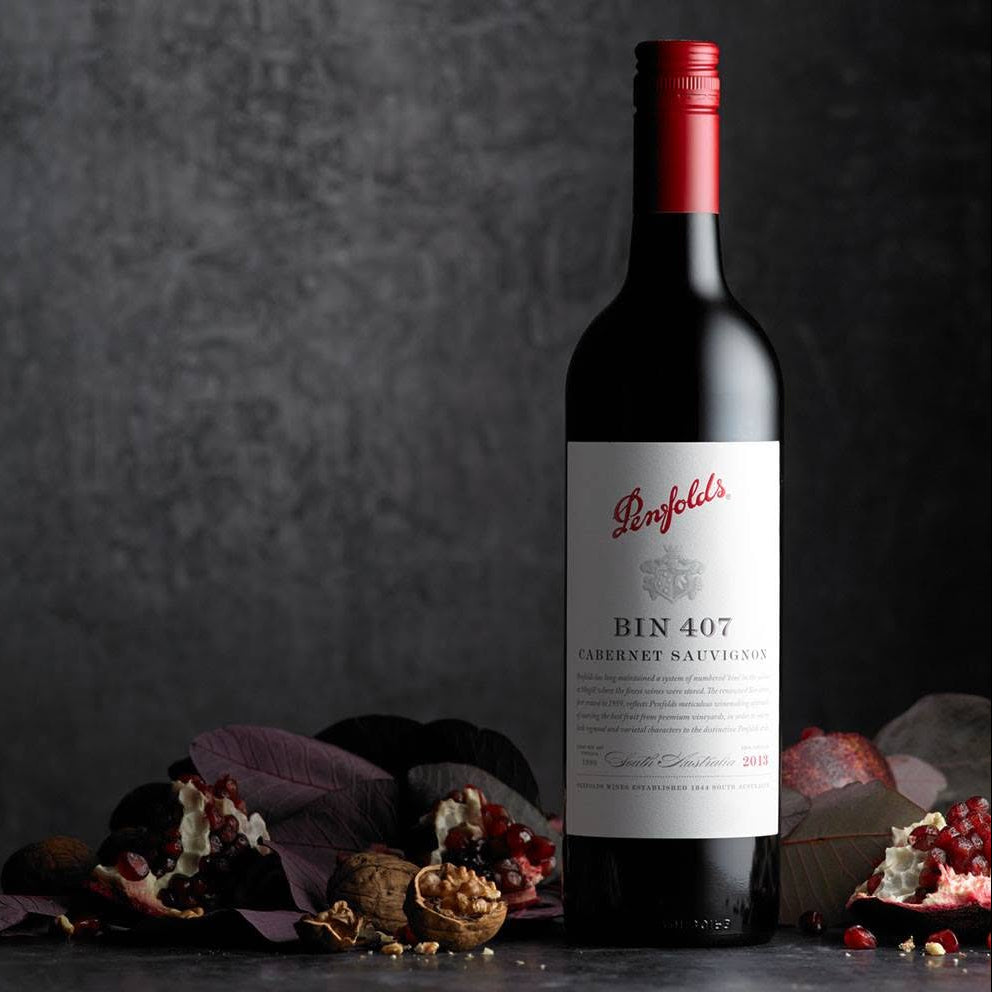 Load image into Gallery viewer, A bottle of Penfold's Bin 407 Cabernet Sauvignon red wine, styled with pomegranate seeds and dried leaves on a dark textured surface against a dark gray background.