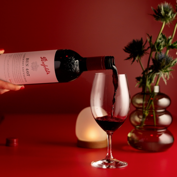 Load image into Gallery viewer, A bottle of Penfolds Bin 407 Cabernet Sauvignon red wine is being poured from a hand into a stemmed wine glass on a red surface, with a red background and decorative thistle flowers in a glass vase.