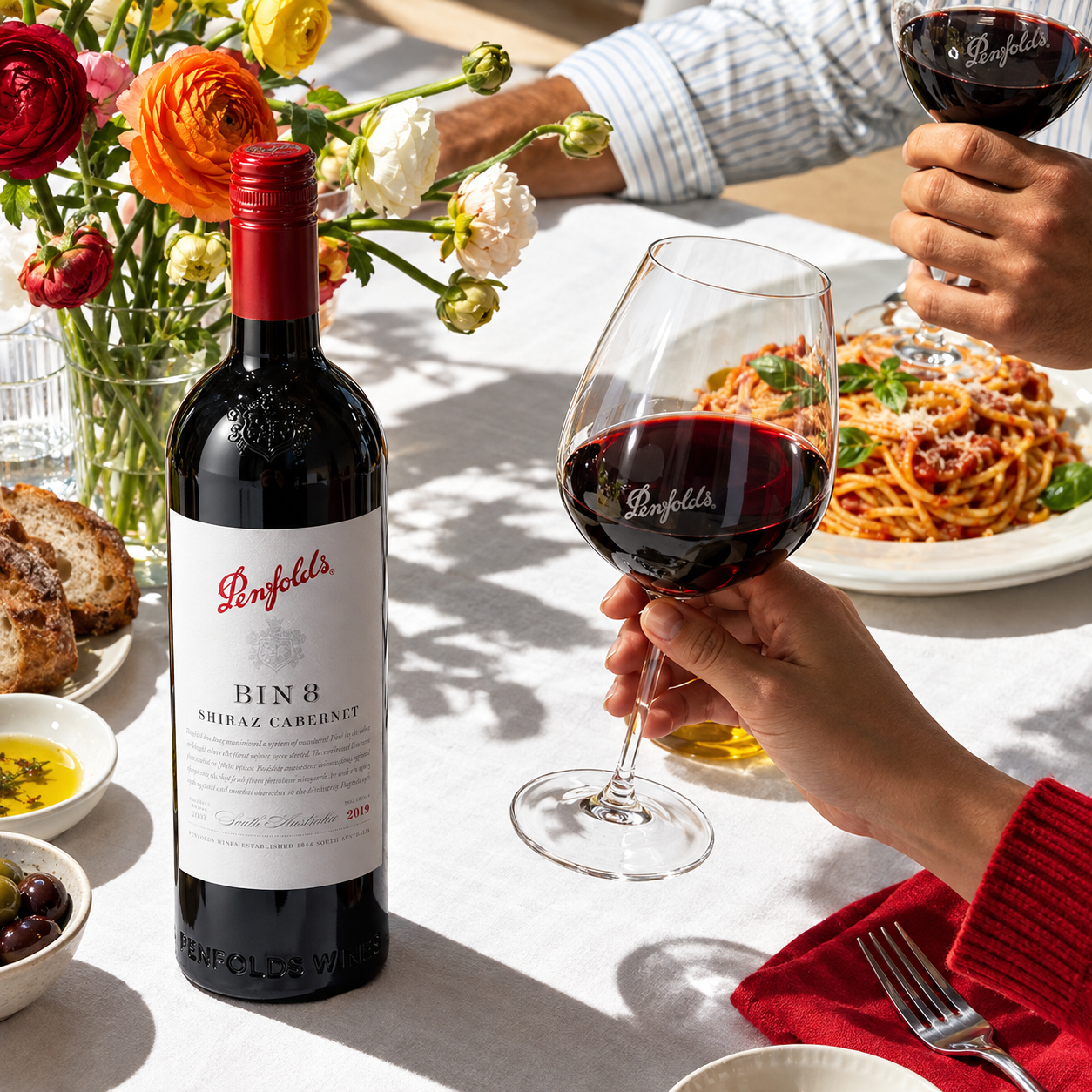 Load image into Gallery viewer, A bottle of Penfolds Bin 8 Shiraz Cabernet red wine and several stemmed wine glasses on a dining table, with people holding glasses of red wine and a plate of spaghetti in the background.