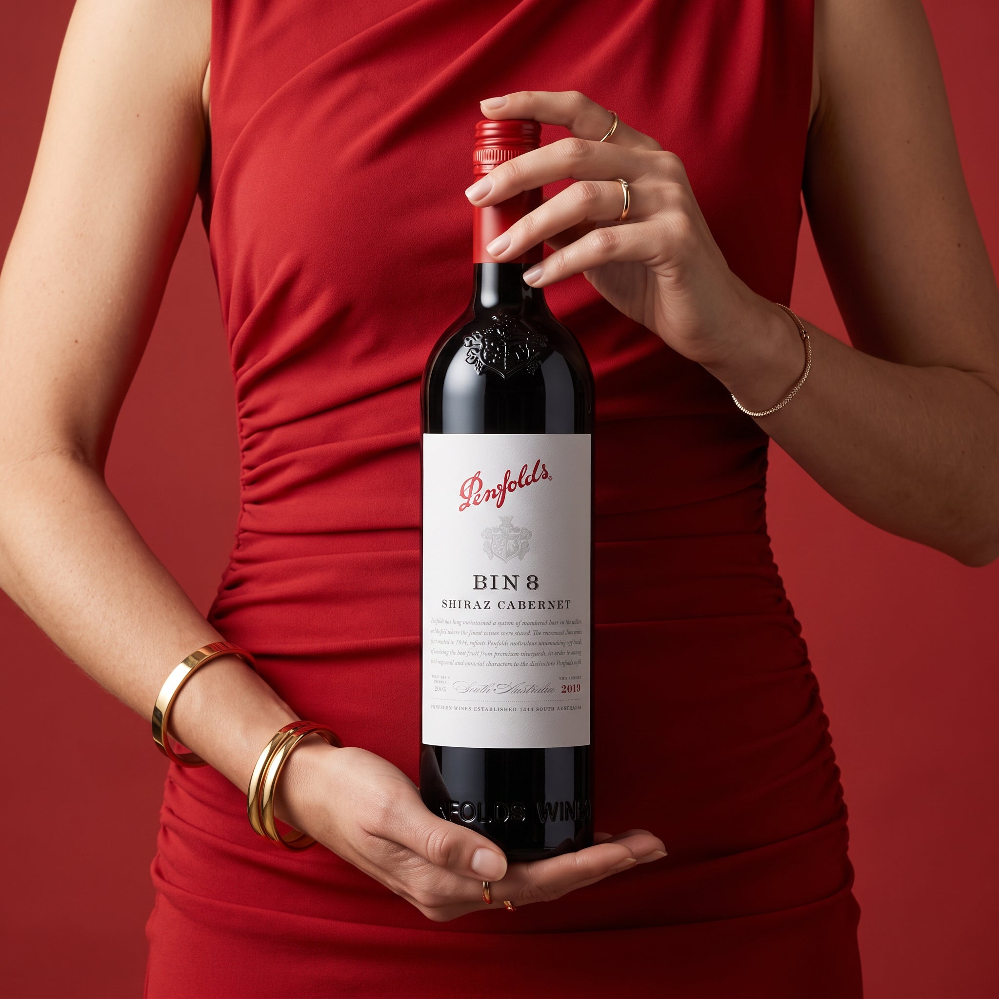 Load image into Gallery viewer, A woman in a red dress holds a bottle of Penfolds Bin 8 Shiraz Cabernet red wine against a solid red background.