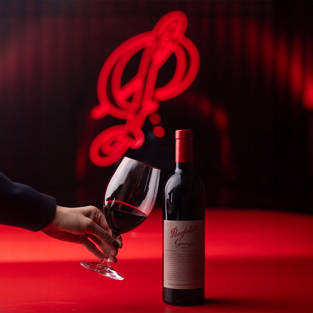 Load image into Gallery viewer, A bottle of Penfolds Grange Red wine and a hand holding a glass of red wine on a red surface, with a red neon-style musical note sign in the background.