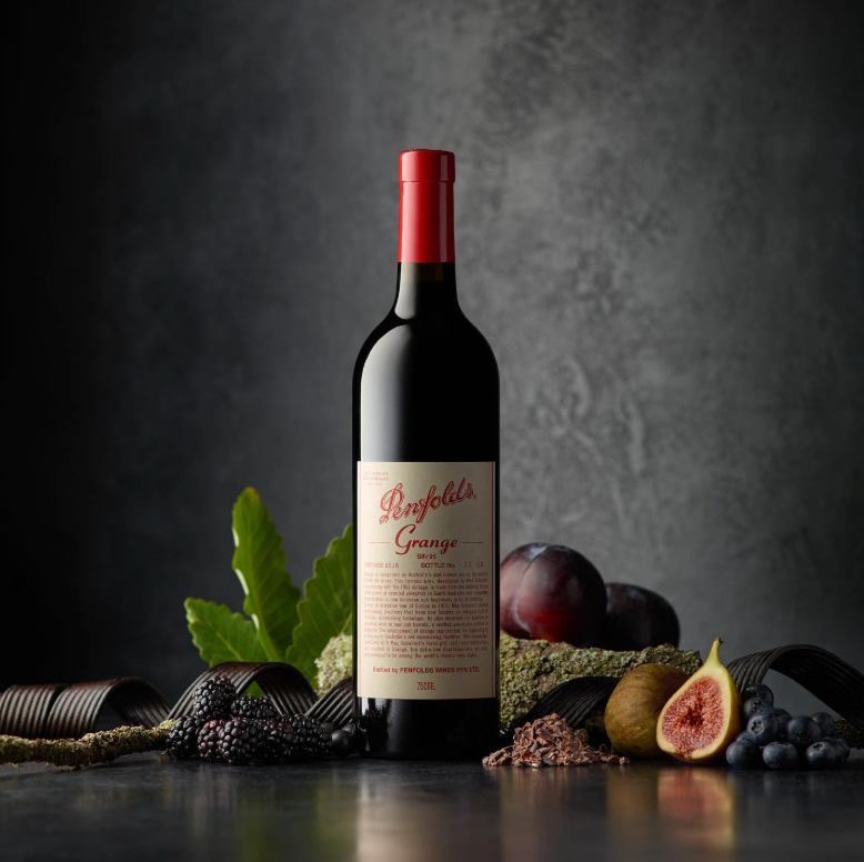 Load image into Gallery viewer, A bottle of Penfolds Grange Shiraz wine stands centred on a dark tabletop, surrounded by food props including plums, figs, blueberries, green leaves, and dried herbs against a textured dark grey background.