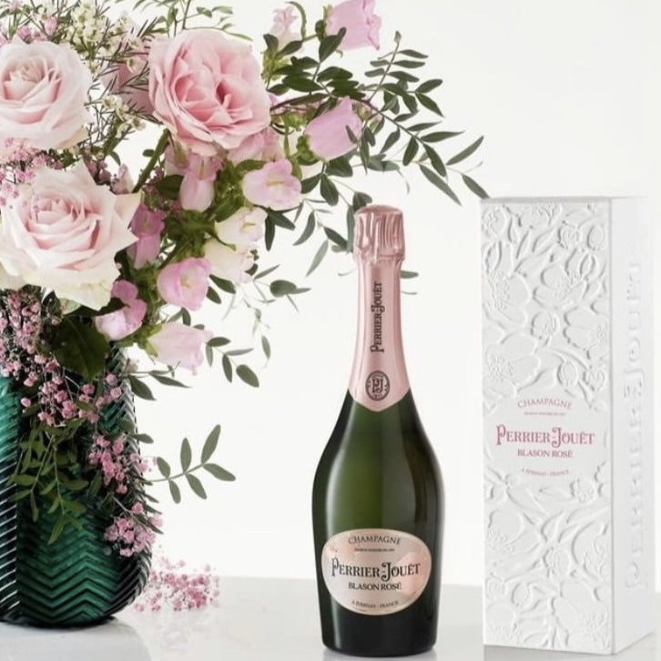 Load image into Gallery viewer, Bottle of champagne with a vase of pink flowers and gift box in the background.