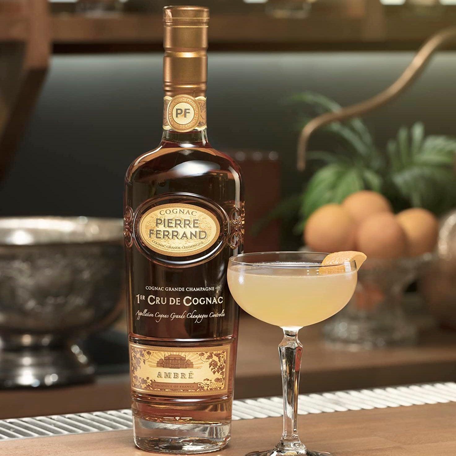 Load image into Gallery viewer, A bottle of Pierre Ferrand Ambre Cognac and cocktail with a lemon wedge on a bar table with a blurry background
