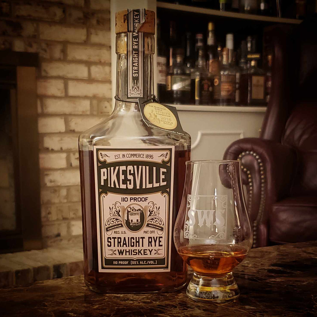 A bottle of Pikesville Whiskey standing on a dark marble table with a tumbler full of whiskey beside it.