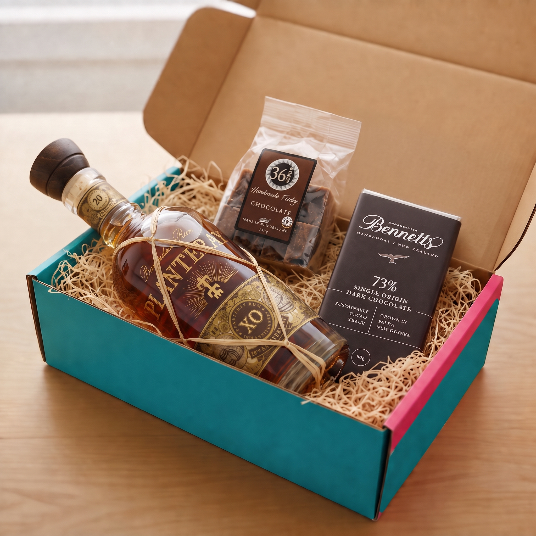 Load image into Gallery viewer, A gift box set on a wooden table containing a bottle of Planteray XO Rum, Bennetts 73% dark chocolate bar, and 36 Degrees chocolate fudge in a turquoise and pink COVE 27 box filled with wood-wool.