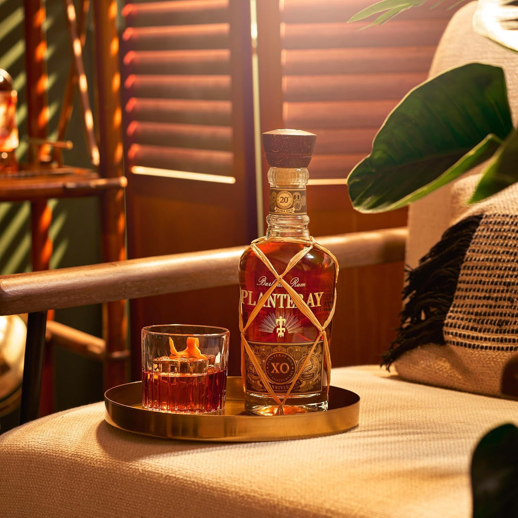 Bottle of Plantation XO rum on a tray with a glass of dark liquid, set against a warm, blurred background.