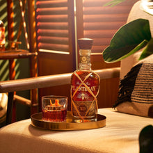 Load image into Gallery viewer, Bottle of Plantation XO rum on a tray with a glass of dark liquid, set against a warm, blurred background.