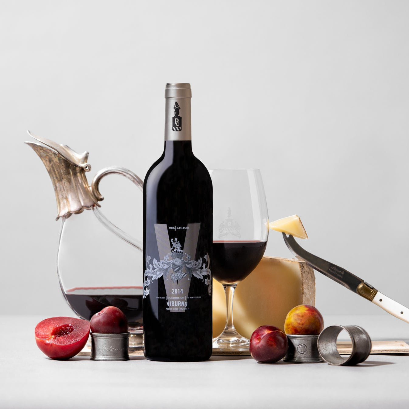 Load image into Gallery viewer, Bottle of Poderi Crisci Viburno red wine with a glass, plums, and a knife on a light gray background