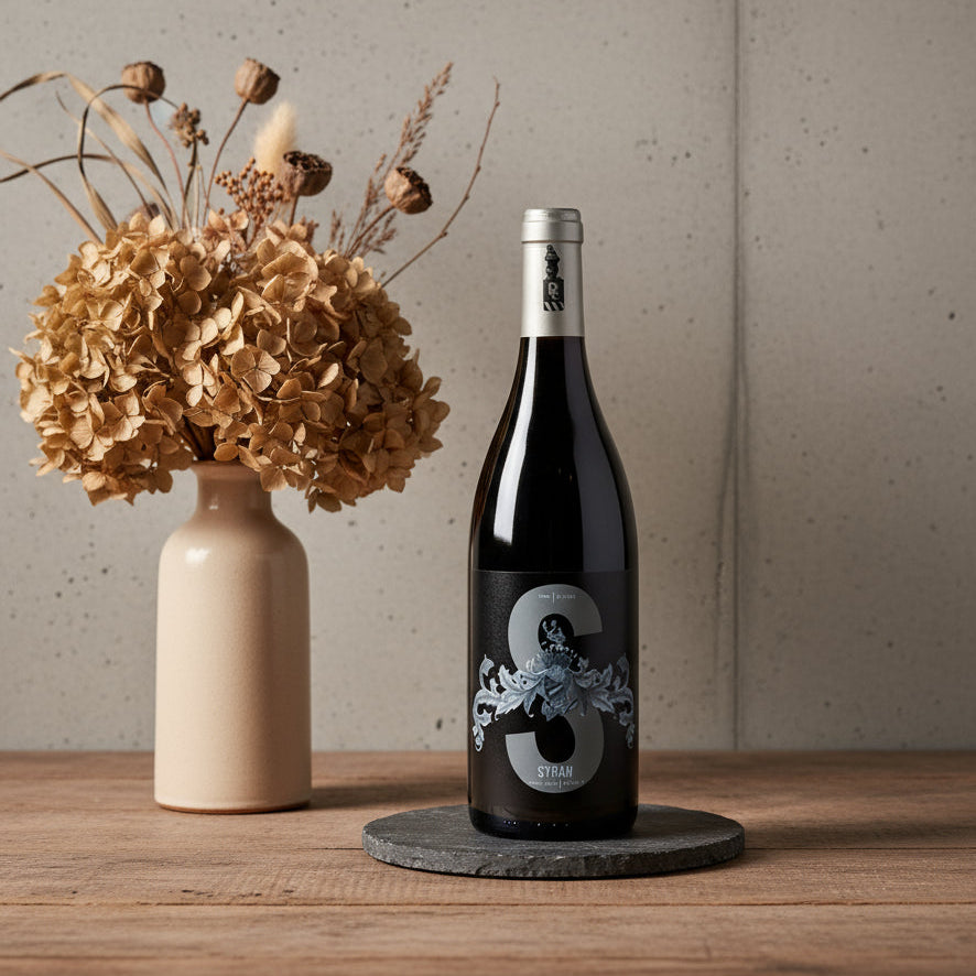 Poderi Crisci Syrah wine bottle with decorative label on a wooden table next to a vase with dried flowers.