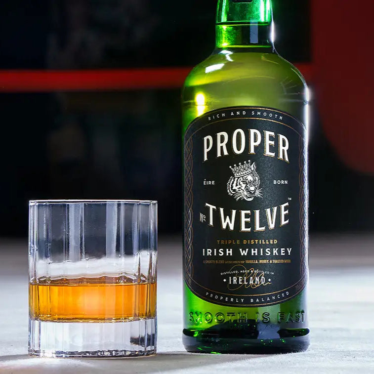 A bottle of Proper Twelve Irish Whiskey  on a white table and a neat glass of whiskey 