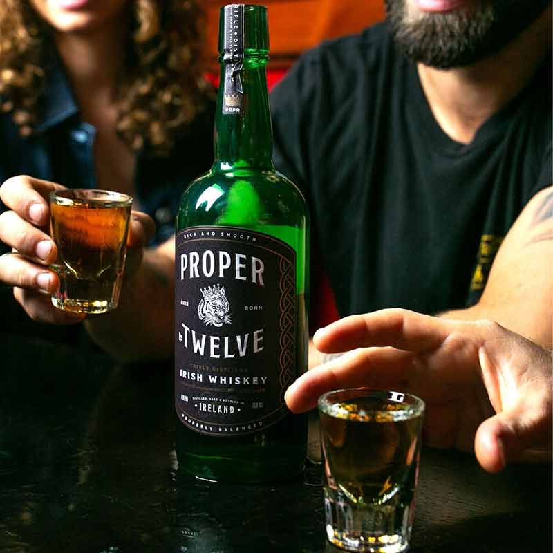 A bottle of Proper Twelve Irish Whiskey  and a couple of people making a cheer with glasses full of whiskey