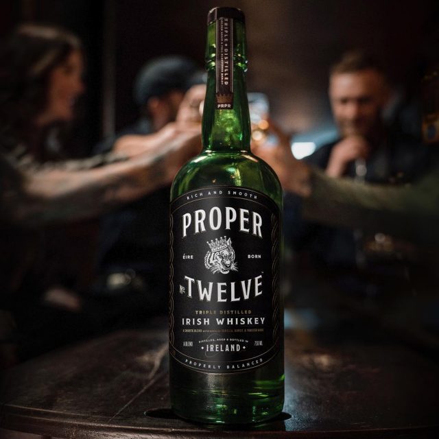 Load image into Gallery viewer, A bottle of Proper No. Twelve Irish Whiskey on a black table with people making a cheer in the background